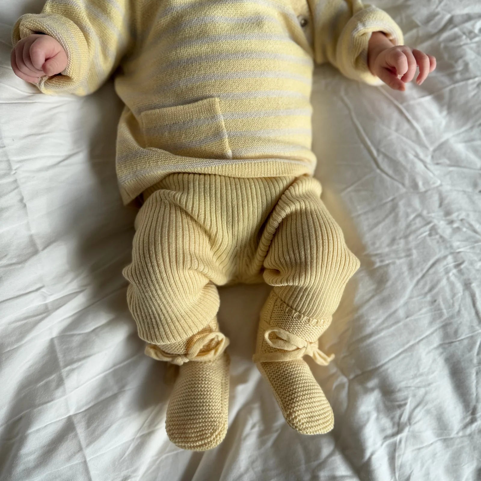 Baby in einem Lille Toni Set aus gestreiftem Merinowolle-Pullover, farblich passender Strickhose und Schühchen in Buttermilk auf weißem Laken.