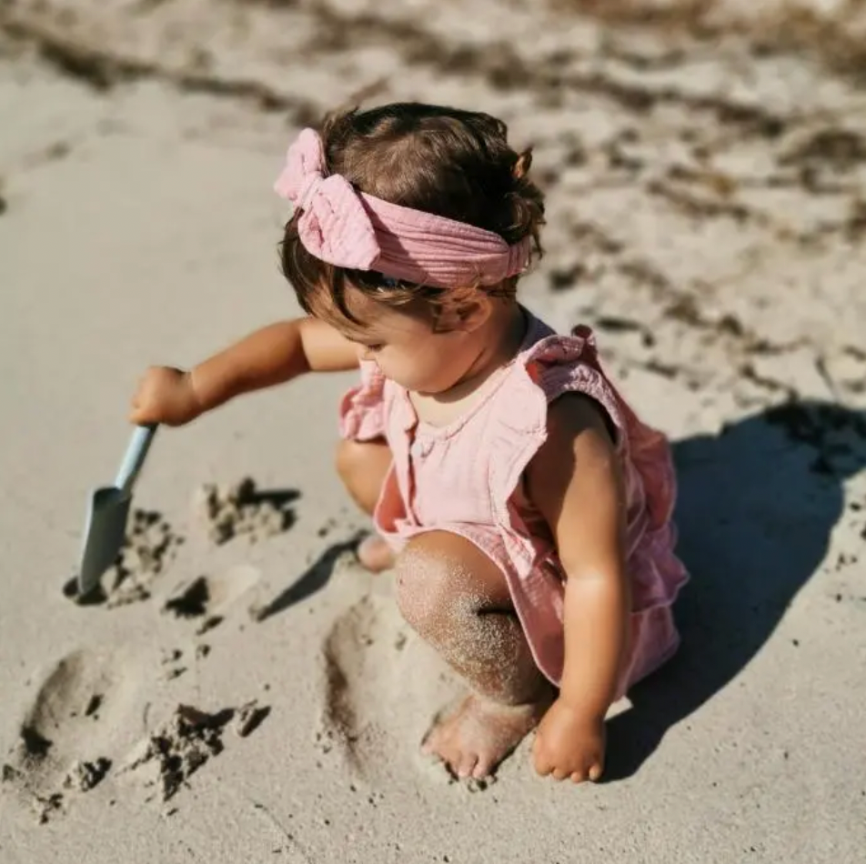 Baby Mädchen im Musselin Top, mit Haarband in rosa spielt mit Sand am Strand 